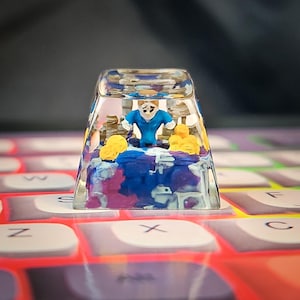May include: A transparent, rectangular keycap featuring a miniature diorama. A small figure in blue stands on a blue, purple, and white base, with yellow figures. The keycap is set on a keyboard.