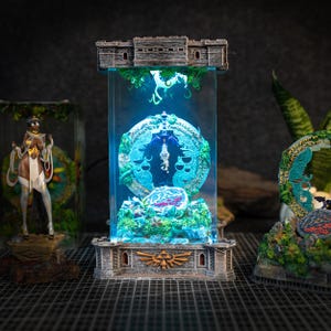 Op de afbeelding: Een glazen vitrine met een blauw licht erin, met een gedetailleerd miniatuur van een draak en een cirkelvormig ontwerp in groen en goud. De vitrine staat op een stenen basis met een decoratief ontwerp.