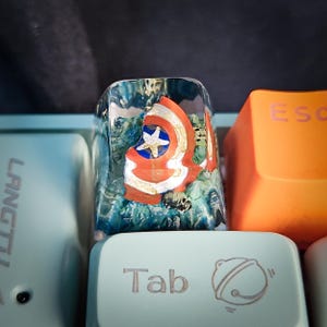 May include: A clear resin keycap with a red, white, and blue Captain America shield design inside. The keycap is on a light blue keyboard with the word "Tab" printed on the key to the right.