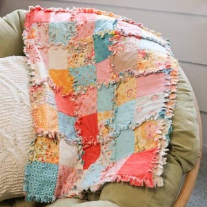 Puede incluir: Un edredón de retazos con borde con volantes, con cuadrados de varias telas florales y estampadas en tonos de rosa, naranja, azul y amarillo. El edredón está sobre una silla verde, mostrando su calidad hecha a mano.