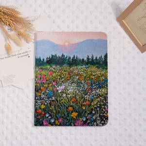 Könnte beinhalten: Ein Notizbuch mit einem farbenfrohen Blumenfeld-Design. Das Cover zeigt ein Gemälde eines Feldes mit Wildblumen, einer Bergkette und einem Sonnenuntergang im Hintergrund. Der Text links lautet "One word then one smile is enough."