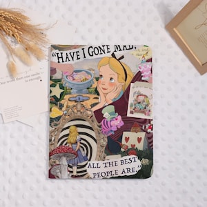 May include: A colorful Alice in Wonderland themed notebook cover. The design features Alice, the Mad Hatter, and other characters from the story. Text includes "HAVE I GONE MAD" and "ALL THE BEST PEOPLE ARE." The cover is a collage of illustrations and text.