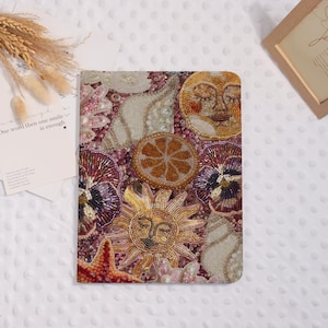 May include: A decorative tablet cover with a beaded design featuring a sun, moon, seashells, and flowers. The cover is primarily pink, with accents of gold, orange, purple, and white. The text on the side says, "One word then one smile is enough."