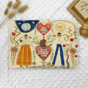 May include: A rectangular art print featuring a folk art style illustration. The artwork depicts two figures, a man and a woman, with a heart between them. The heart contains the text "I WILL NEVER DECEIVE YOU" and "DO NOT DECEIVE ME". The background includes a moon, sun, and floral elements.