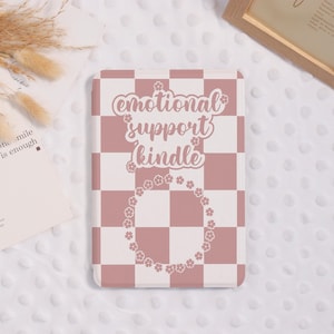 May include: A Kindle case with a pink and white checkered pattern. The case features the text "emotional support kindle" in a cursive font, surrounded by a floral wreath. The case is designed to protect and personalize a Kindle device.