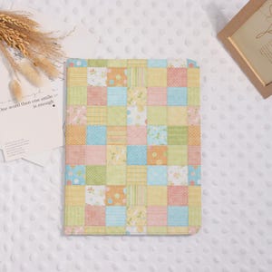 May include: A colorful patchwork pattern notebook cover in pastel shades of yellow, pink, green, and blue. The cover is made of squares with floral and polka dot designs. The notebook is lying on a white, textured surface.