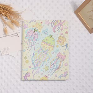 May include: A pastel yellow notebook featuring a whimsical design of cartoon jellyfish, stars, and cat-like creatures. The cover has a soft, dreamy aesthetic with a variety of colors, including pink, blue, and green.