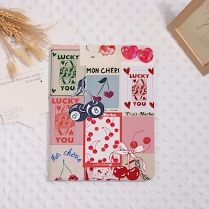 May include: A decorative notebook with a collage design featuring cherries, playing cards, and the phrases "Lucky You," "Mon Chéri," and "Ma chérie." The color palette includes pink, red, and white.
