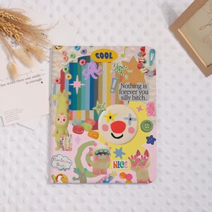 May include: A rectangular notebook cover featuring a collage of colorful graphics, including a clown face, a giraffe, a monster, stars, and text that reads "COOL," "Nothing is forever you silly bitch," "Fun in the Sun," and "Nice" on a pastel background.