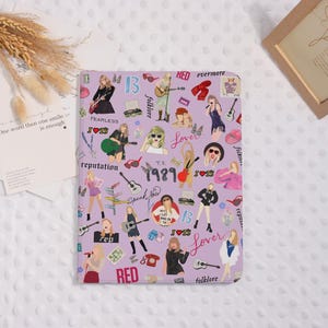 May include: A lilac notebook adorned with illustrations of a singer, guitars, and album titles. The notebook showcases a repeating pattern of the singer's image, musical instruments, and album names such as "Lover" and "Red".
