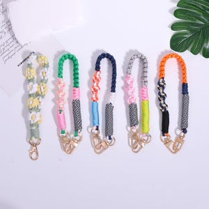 May include: Five colorful keychains with gold-colored clasps. The keychains feature braided and woven designs in various colors, including green, pink, blue, orange, and gray. One keychain has a daisy pattern.
