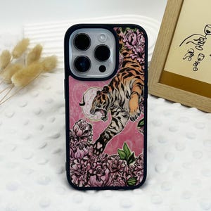 May include: Black phone case with a pink background featuring a tiger illustration. The tiger is orange and black, surrounded by pink flowers and green leaves. The case has a black border.
