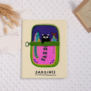 May include: A beige notebook featuring a whimsical illustration of a sardine can. The can is green and pink, with a black cat peeking out between two colorful fish. The word "SARDINES" is printed on the cover.