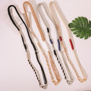 May include: An assortment of six woven eyeglass straps in various colors, including black, tan, gray, and cream. Each strap features a braided design and a metal clasp for attaching to glasses. The straps are displayed on a light-colored surface.