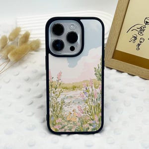 Peut inclure: Une coque de téléphone noire avec un fond blanc et une illustration colorée d'un étang avec des canards et des fleurs. La coque de téléphone a un protège-objectif d'appareil photo transparent.