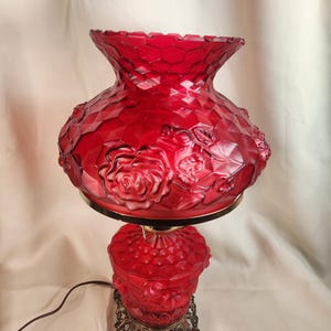 May include: A vintage red glass table lamp with a floral rose design. The lamp has a textured shade and base, with a decorative brass-coloured metal stand. The lamp is plugged in and ready to use.