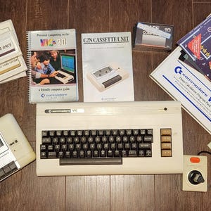 May include: A vintage Commodore VIC-20 computer with accessories, including a cassette unit, game cartridges, manuals, and a joystick. The computer is beige with a black keyboard. The items are arranged on a wooden surface.