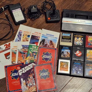 Könnte beinhalten: Eine Sammlung von Vintage Atari Videospielkonsolen, Controllern, Spielkassetten und Katalogen. Die Konsole ist schwarz mit einem silbernen Streifen. Spielkassetten enthalten Titel wie "Smurf" und "Asteroids".