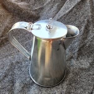 Może przedstawiać: Mały, błyszczący, srebrny metalowy dzbanek z uchwytem i pokrywką. Dzbanek ma dzióbek do nalewania i pokrywę na zawiasach z małą pętlą do podnoszenia. Dzbanek jest prawdopodobnie używany do serwowania płynów.