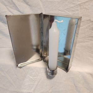 Puede incluir: Un candelero de metal plateado con una vela blanca en su interior. El candelero tiene una bisagra y se abre para revelar un pequeño compartimento vacío.