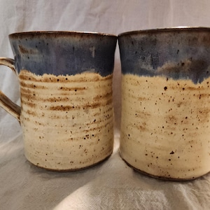 Puede incluir: Dos tazas de cerámica hechas a mano. Una tiene asa, la otra es un vaso. Ambas tienen un borde azul, un cuerpo beige moteado y rayas horizontales marrones. Las tazas están sobre una tela blanca.