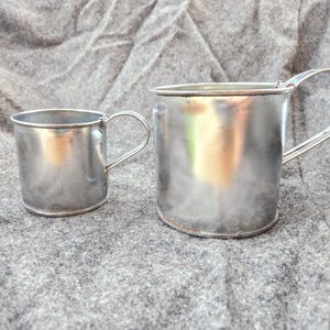 May include: Two small, shiny, silver metal mugs with curved handles sit on a gray, textured surface. The mugs are different sizes, with the larger one on the right. The mugs have a simple, classic design.