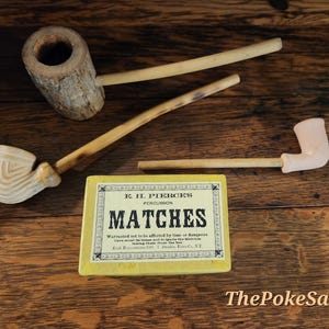 Puede incluir: Tres pipas de fumar y una caja rectangular amarilla de cerillas E.H. Pierce's Percussion. Una pipa tiene un hornillo con textura de corteza, otra presenta un hornillo blanco acanalado y la tercera tiene un hornillo blanco liso con un largo tallo de madera.