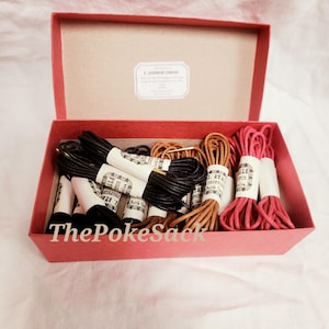 Puede incluir: Una caja roja llena de cordones de diferentes colores. Los cordones son negros, marrones y rosas, están empaquetados y etiquetados con papel blanco. La caja tiene una etiqueta dentro de la tapa y el texto "ThePokeSack" en la parte delantera.