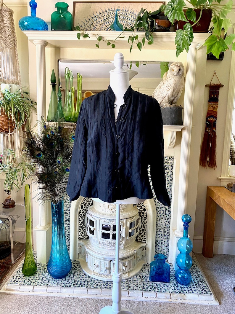 Puede incluir: Una camisa negra de manga larga con botones y cuello mandar&iacute;n. La camisa est&aacute; en un maniqu&iacute; blanco frente a una chimenea.