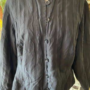 Puede incluir: Una camisa negra de manga larga con cuello mandar&iacute;n y pliegues verticales. La camisa tiene una serie de botones negros en la parte delantera.