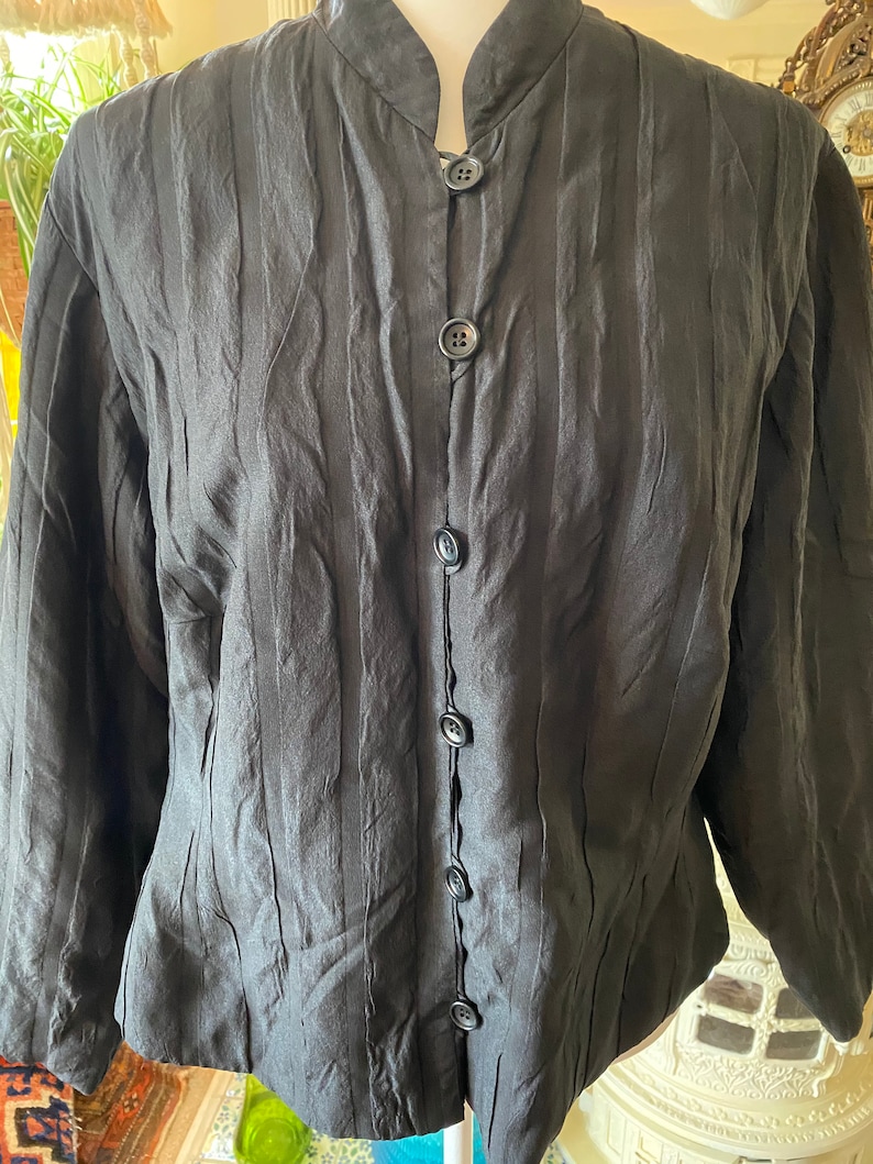 Puede incluir: Una camisa negra de manga larga con cuello mandar&iacute;n y pliegues verticales. La camisa tiene una serie de botones negros en la parte delantera.