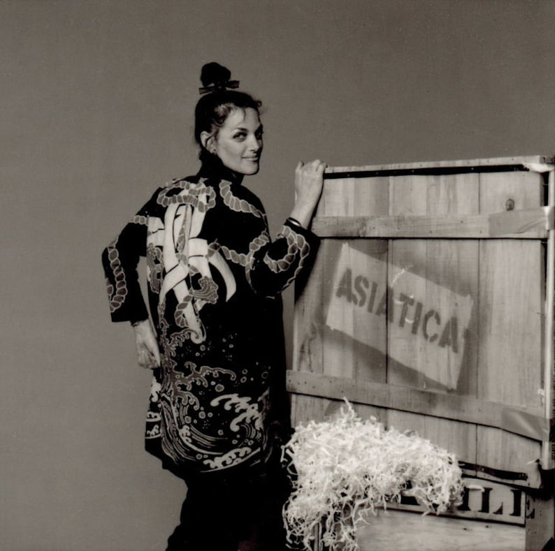 Puede incluir: Una mujer con una chaqueta de estilo kimono con estampado en blanco y negro se encuentra frente a una caja de madera con la palabra "ASIATICA" impresa en ella. La caja est&aacute; llena de material de embalaje blanco.