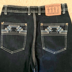 May include: Back view of a pair of dark blue denim jeans with a white stitch detail. The back pockets have a geometric design embroidered in white and blue thread. A brown leather patch with the text "FIORUCCI" and "SUPER JEANS" is sewn on the right back pocket.
