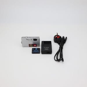 May include: A silver digital camera with a black battery pack, charger, and power cord. The camera has a lens and a flash. The battery pack has the text "Panasonic" and "Battery Pack". The charger has the text "LUMIX".