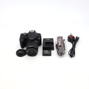 Op de afbeelding: Een zwarte Canon EOS 1300D DSLR-camera met een lens, lensdop, batterij, oplader en riem. De camera heeft een zwarte body en een zilveren lens. De accessoires omvatten een oplader, batterij en een zwarte en rode riem.