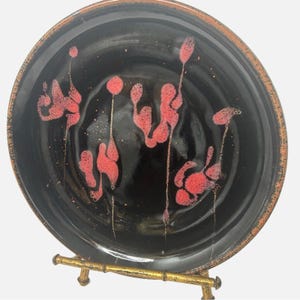 Puede incluir: Un plato de cerámica negro con un esmalte rojo y dorado. El plato tiene un borde elevado y está decorado con un patrón de remolinos rojos y dorados. El plato está sobre un soporte dorado.