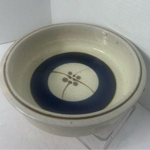 Può includere: Una ciotola rotonda in ceramica con smalto bianco sporco maculato. L'interno presenta un'ampia fascia circolare blu scuro che circonda un cerchio centrale color crema decorato con un motivo floreale marrone a quattro petali e steli curvi.