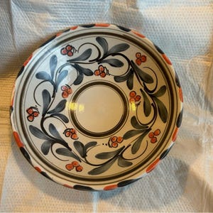 Puede incluir: Un cuenco decorativo de cerámica con interior blanco y borde ancho. El cuenco presenta un diseño pintado a mano de hojas grises, bayas rojas y detalles en negro y naranja. El centro del cuenco tiene un diseño circular.