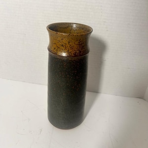 May include: A cylindrical ceramic vase featuring a dark charcoal grey base with a speckled texture that transitions into a distinct, wider, golden-brown rim. The vessel has a smooth, rounded opening and a straight, vertical body profile.