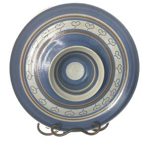 Pode incluir: Um prato redondo de cerâmica com uma tigela central rebaixada, decorado com faixas concêntricas em azul, creme e castanho. Um anel de pequenos corações pretos desenhados à mão rodeia o centro sobre um acabamento liso e vidrado.