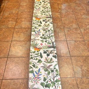 May include: A long, rectangular fabric table runner featuring a white background printed with a repeating botanical pattern of green ferns, leafy trees, colorful orange and yellow butterflies, and small red and blue floral accents throughout.