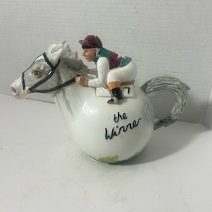 May include: A white ceramic teapot shaped like a horse with a jockey riding on top. The jockey wears a maroon cap and green vest. The teapot has the words "the Winner" written on it.
