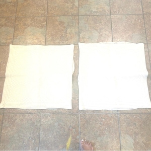 May include: Two off-white square pillowcases are laid out on a tiled floor. One pillowcase has a quilted pattern, while the other is plain. The pillowcases are made of a soft, possibly cotton, material.