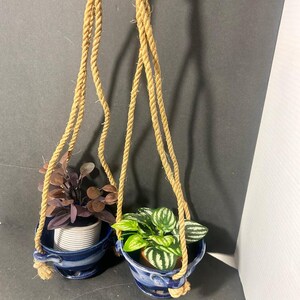 May include: Two blue ceramic hanging planters with integrated saucers and side handles, suspended by thick tan jute ropes. One planter holds a plant with dark purple leaves, while the other contains a plant with green and white striped leaves.