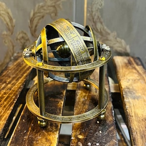 Puede incluir: Una esfera armilar de latón con símbolos del zodiaco y texto. Se encuentra sobre un soporte a juego. El fondo presenta un papel pintado con motivos y una superficie de madera. El diámetro es de aproximadamente 15 cm.