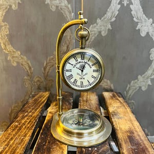 Puede incluir: Un reloj de escritorio de latón de estilo vintage con diseño de reloj de bolsillo. La esfera del reloj indica "Victoria London 1876" con números romanos. Una brújula está en la base. El reloj está soportado por un soporte de latón curvado.