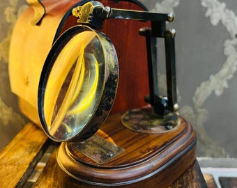 May include: A vintage-style magnifying glass with a wooden base and a brass frame. The magnifying glass is positioned in front of a leather case. The base is oval-shaped and made of dark wood. The magnifying glass is round and has a clear lens.