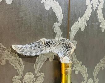 May include: A clear crystal cane with a detailed, bird-shaped handle. The handle sits atop a gold-colored metal collar and a black wooden shaft. The cane is a decorative accessory.