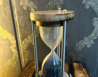 Op de afbeelding: Een houten zandloper met een helder glazen midden gevuld met donkerblauw zand. De zandloper wordt ondersteund door zilveren metalen staven en heeft een houten boven- en onderkant. De houten basis rust op een houten oppervlak.