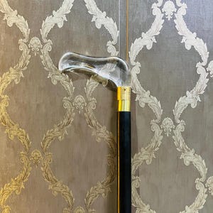 May include: A clear, curved glass handle cane with a gold-colored metal band and a black wooden shaft. The cane is a classic walking aid, combining functionality with an elegant design.